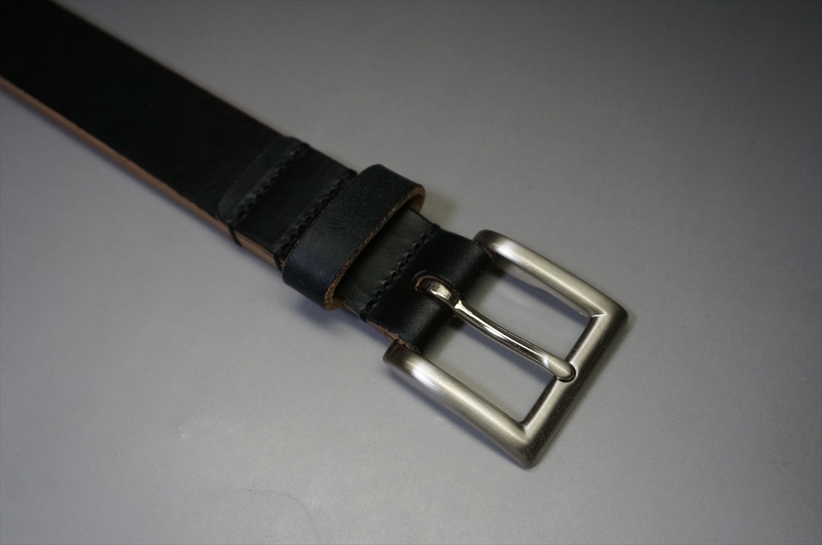 Black leather belt with a silver rectangular buckle lying on a gray surface, close-up of the strap looped through the buckle edge.”