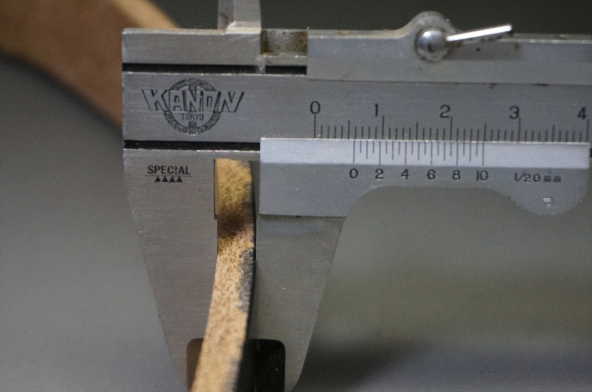 Close-up of a metal caliper measuring a wooden strip, showing the ruler scale around 2 cm.