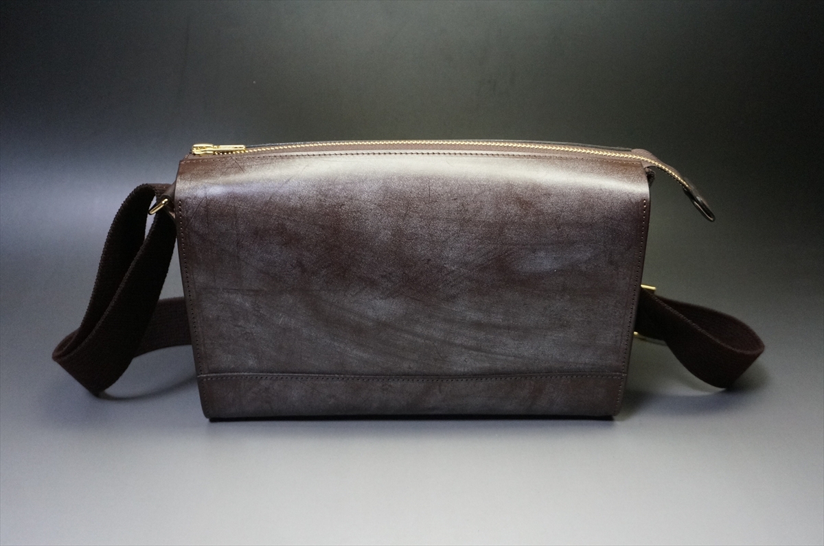 Brown leather crossbody bag with a gold zipper, shown on a gray gradient surface near the camera edge.