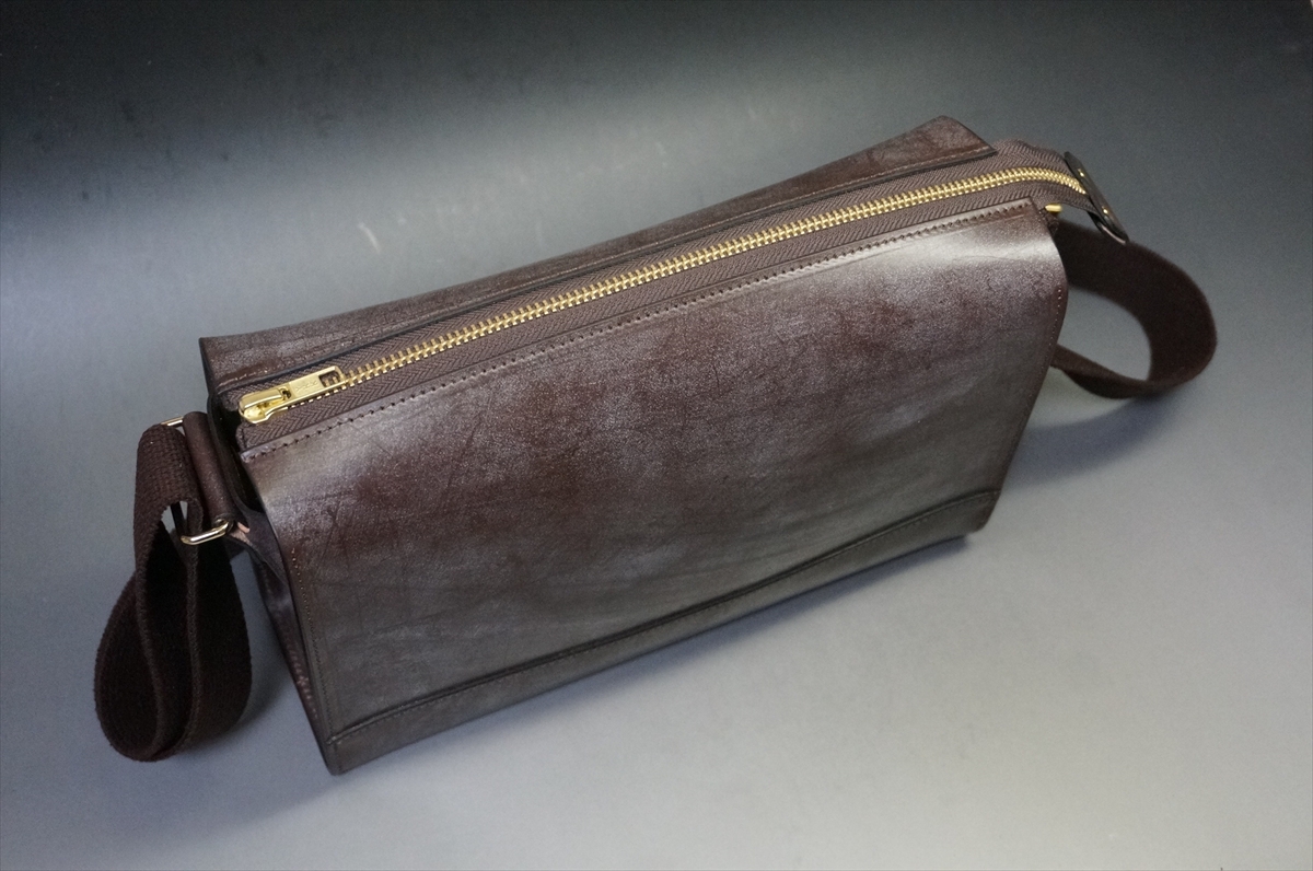 Brown leather crossbody bag with a gold zipper and dark adjustable strap on a gray surface.