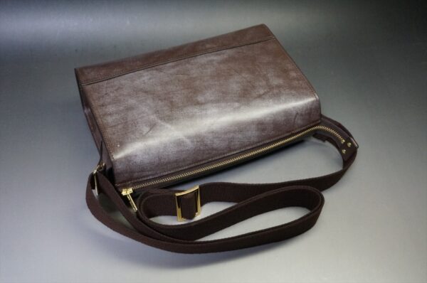 Brown leather crossbody bag with a gold zipper and an adjustable dark fabric strap on a neutral surface.