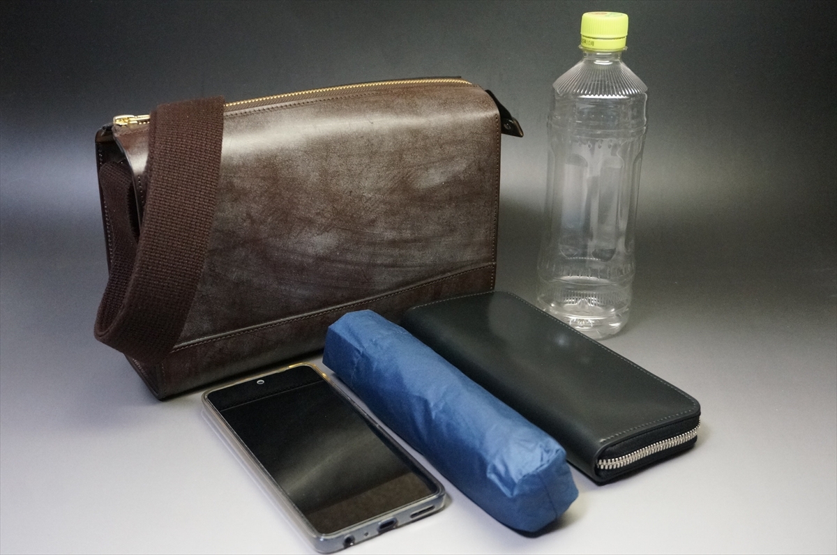 Brown leather messenger bag with strap, placed behind other items on a gray surface. One zipped compartment visible.