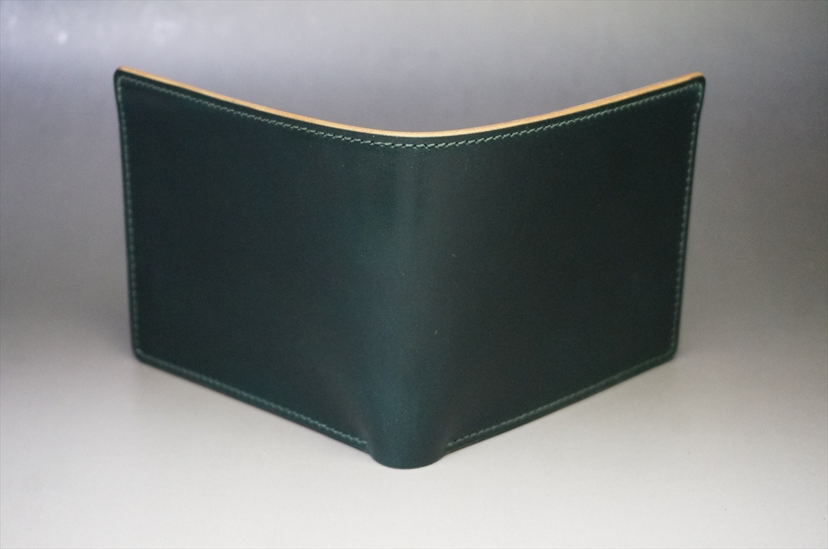 Dark green leather bi-fold wallet with tan edging, standing upright on a gray surface with visible white stitching along the seams.