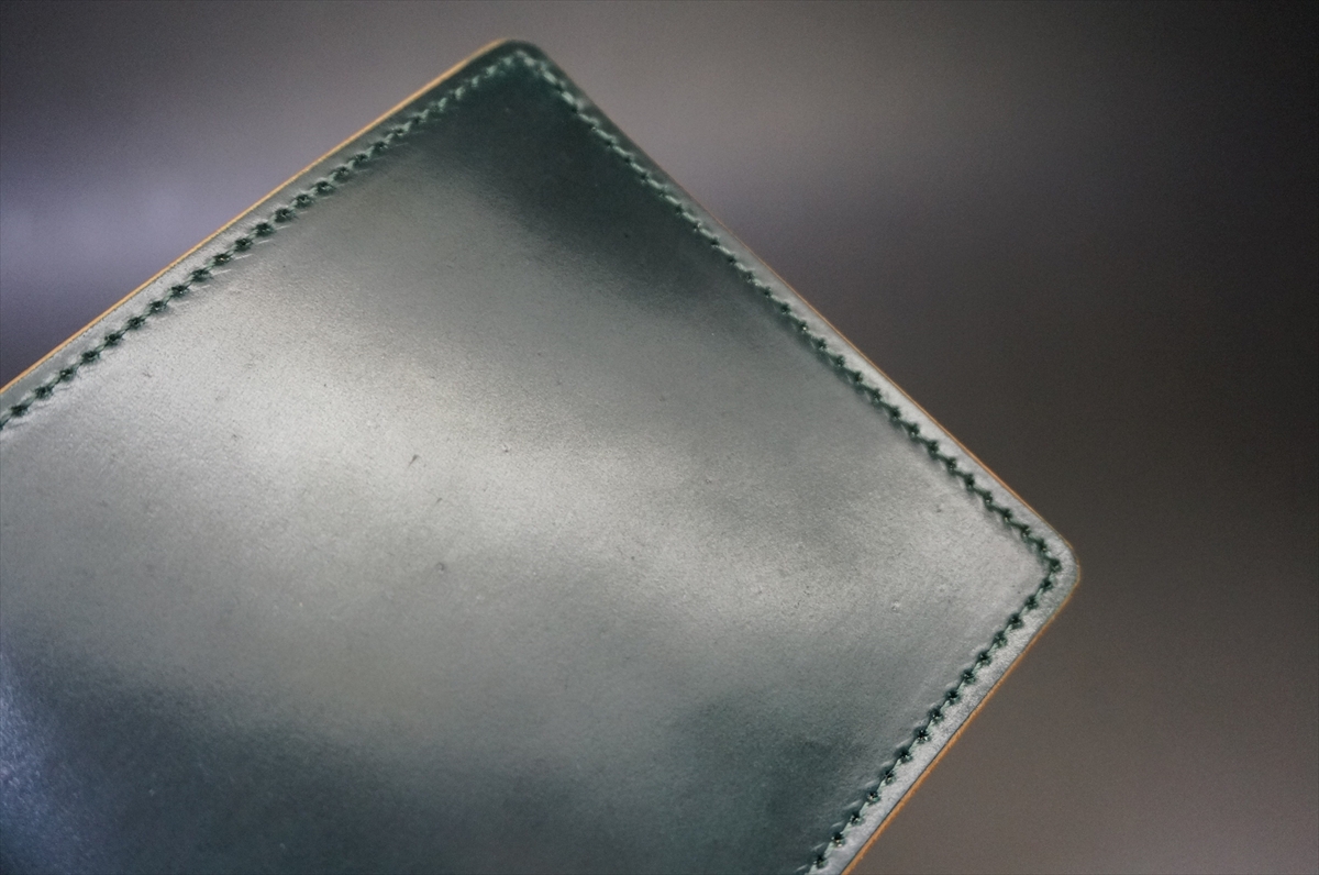 Close-up of a dark green leather wallet corner with even stitching along the edge","Detail of green leather edge and stitching on a wallet or card holder","Angle shot of a green leather surface with stitched border, blurred background","Green leather item with visible stitching, likely a wallet or passport cover","Stitched green leather corner with tan edge trim