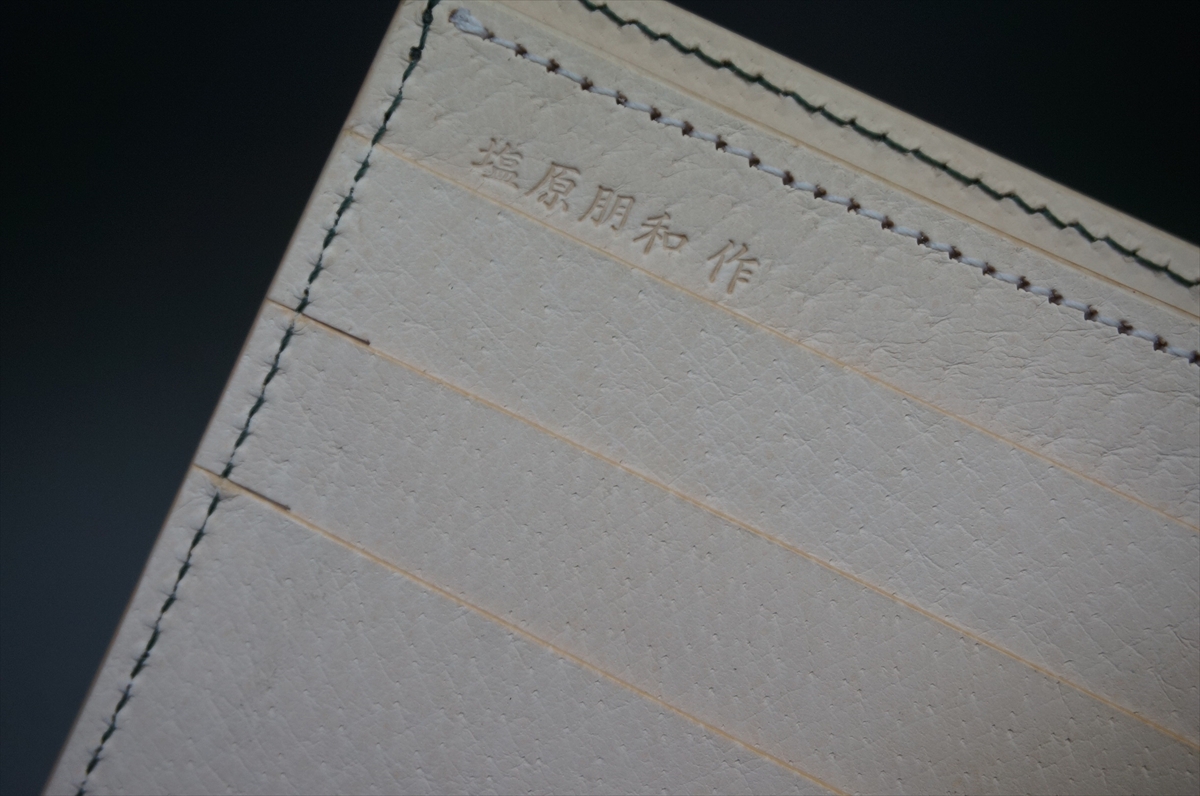 Close-up of a white textured notebook page with pale horizontal lines and green stitching along the edge; embossed Japanese characters near the top.