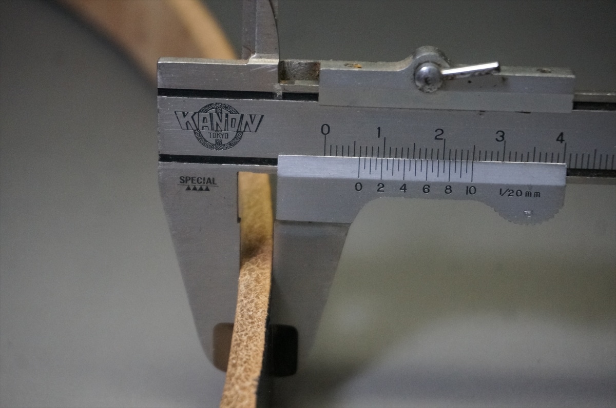 Close-up of a metal caliper measuring a brown leather strap, the scale showing about 2.5 cm from the jaws together with metric markings nearby.