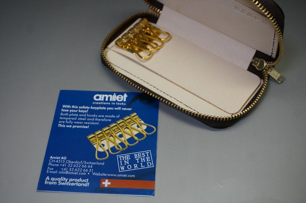 Blue Amiet card advertising a gold key hook set, placed beside an open beige zipper case on a gray surface.