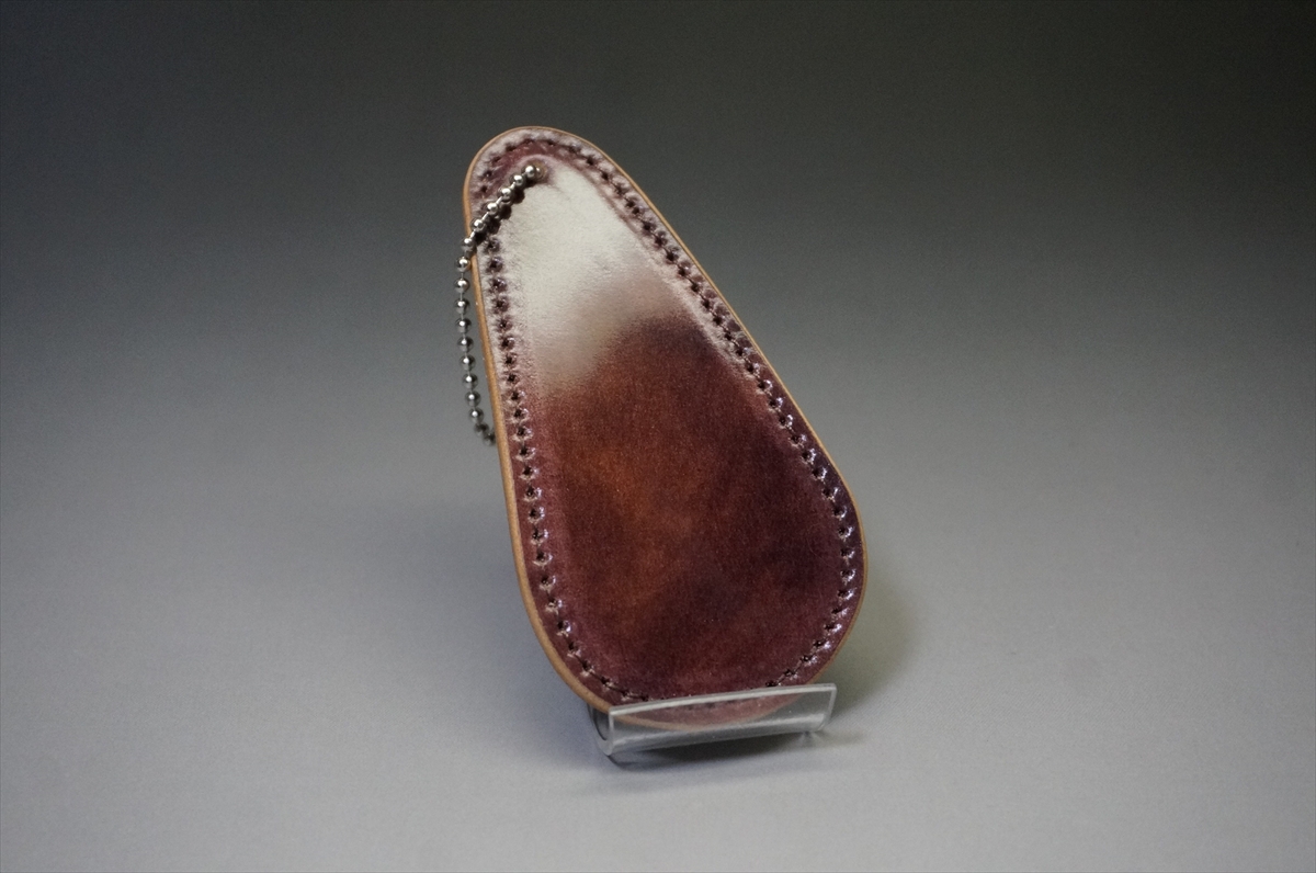 Teardrop-shaped brown leather pouch with stitched edges and a metal ball chain, displayed on a small stand.