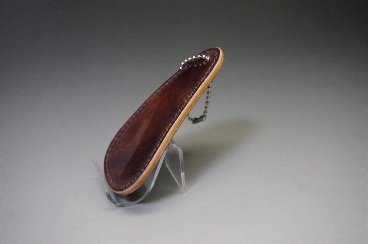 Brown leather key holder with a small metal ball chain, displayed on a clear stand against a neutral background.