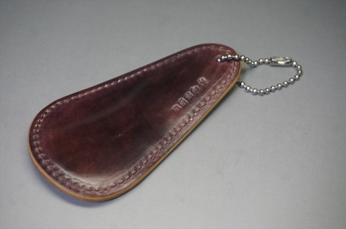 Brown leather key fob with stitching and a small metal ball chain attached, lying on a gray surface. It appears to be a compact leather key case or sheath with a keyring.