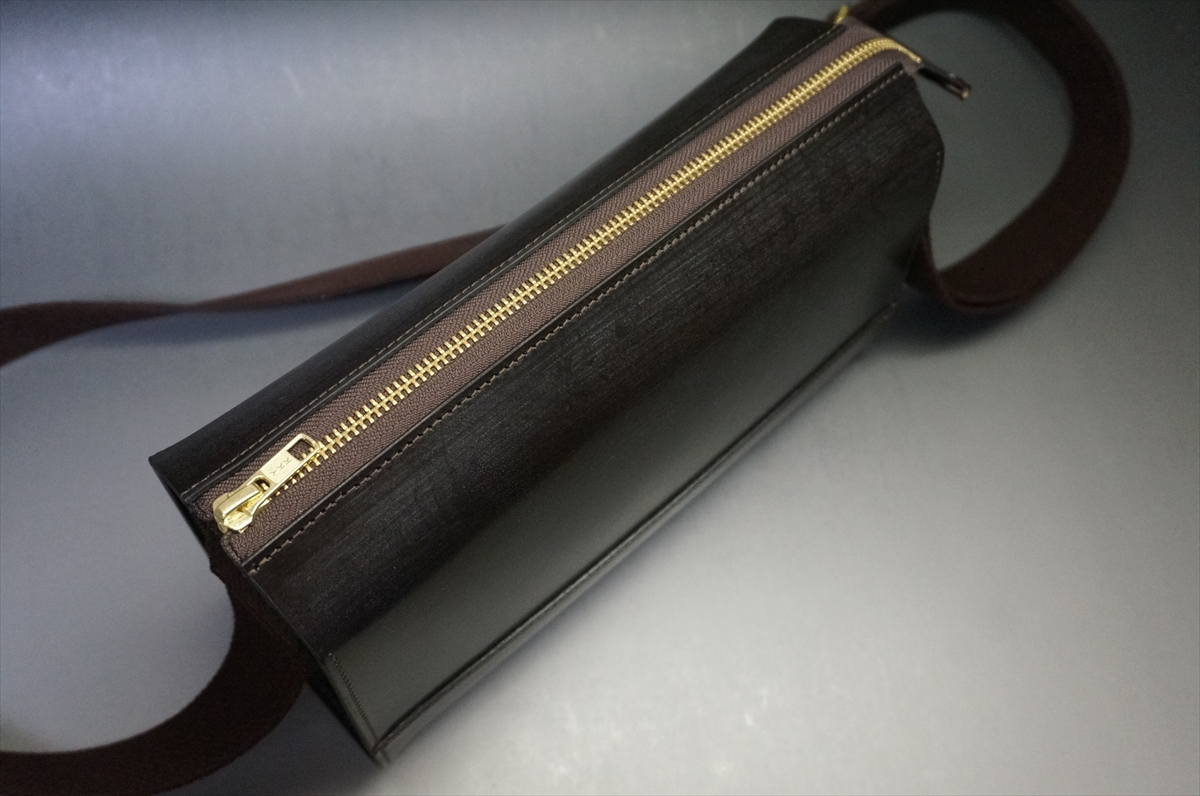 Black rectangular crossbody bag with a gold zipper along the top and a brown strap tails off to the side.