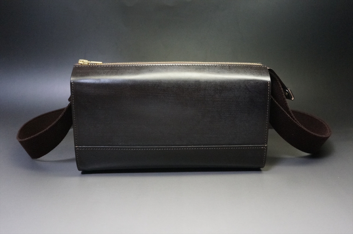 Black leather camera bag with a brown adjustable strap, zipper closure, placed on a neutral surface against a dark backdrop.