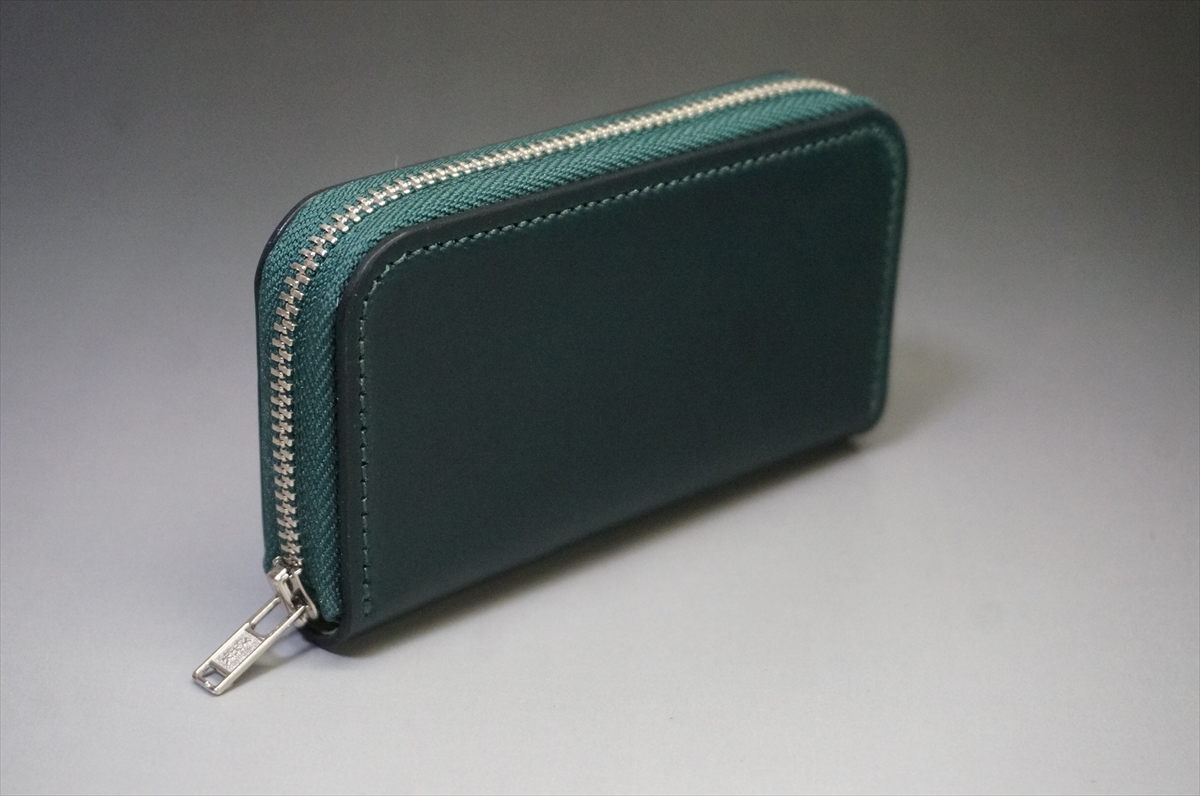 Teal zippered wallet made of leather on a light gray background, showing a silver zipper along the edge.