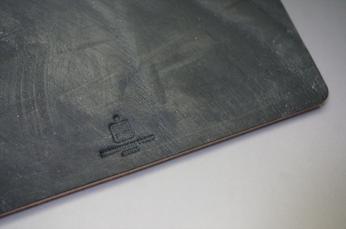 Close-up of a dark, worn metal plate with a small embossed square logo and faint, unreadable text near the edge.