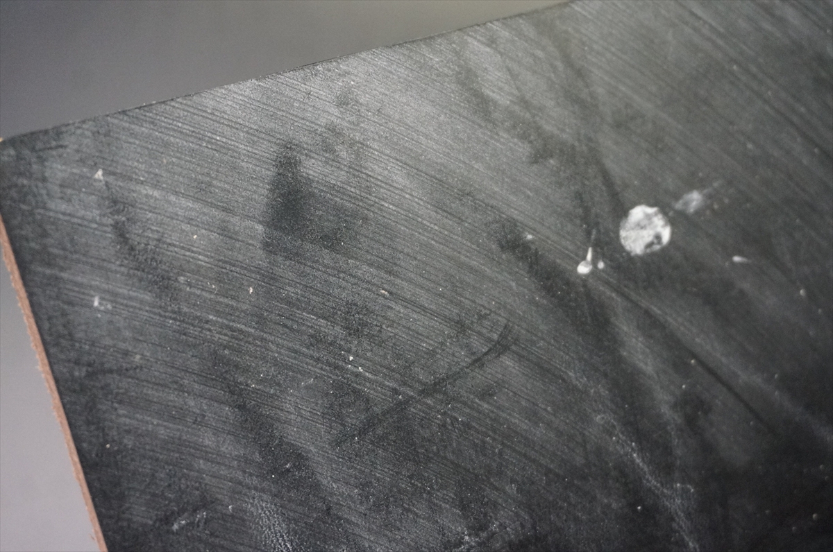 Close-up of a dark, brushed surface (likely wood or panel) with visible grain, scratches, and a few white paint specks.