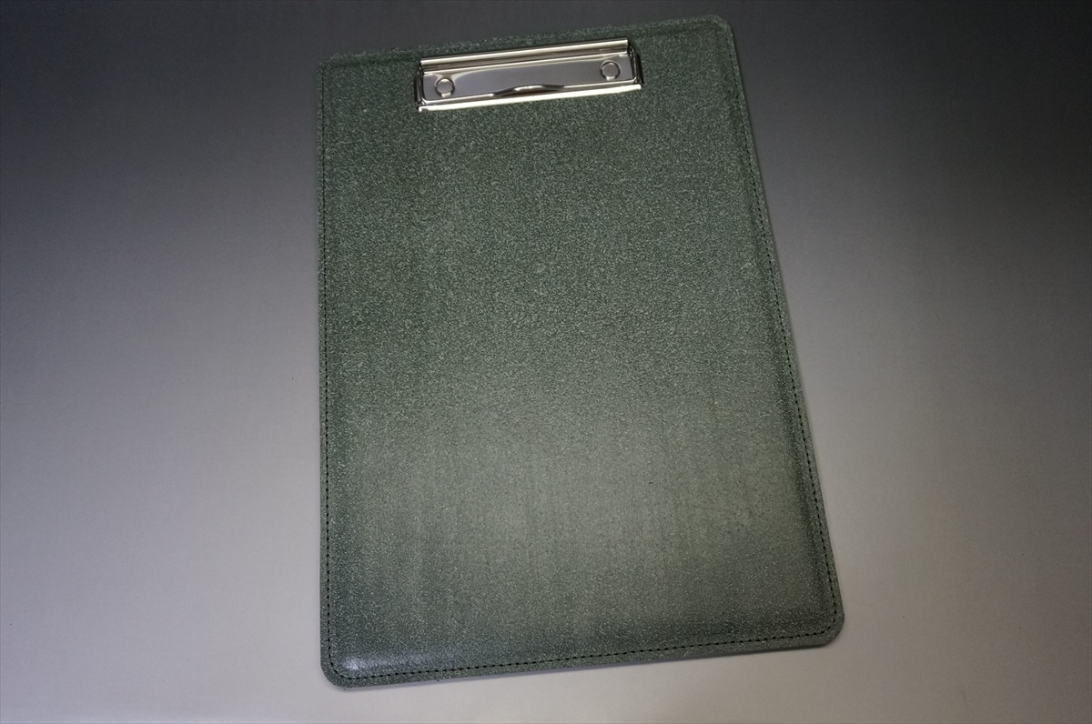 Dark green clipboard with a silver metal clip at the top, viewed from above on a gray surface.