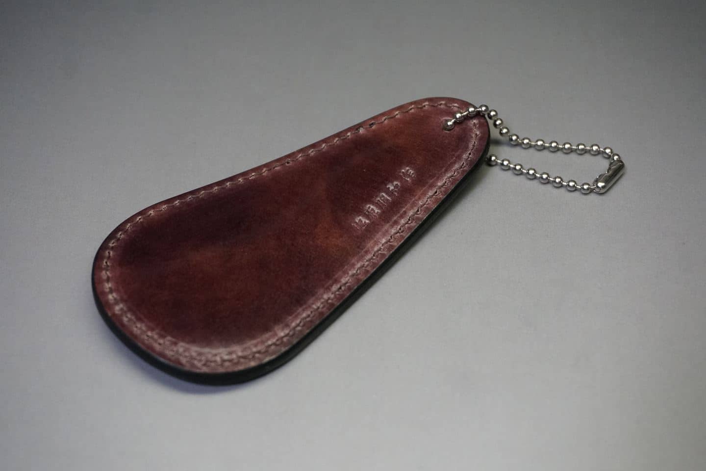 Brown leather key fob pouch with stitched edges and a silver ball-chain attached to one end, lying on a gray surface.