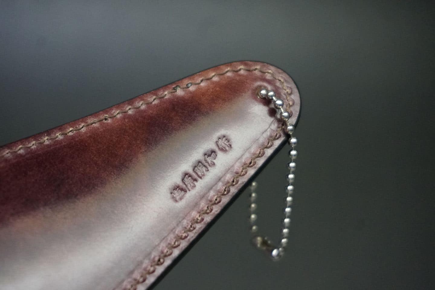 Close-up of a shiny brown leather sheath with decorative stitching along the edge and a small silver ball-chain attached at the end.